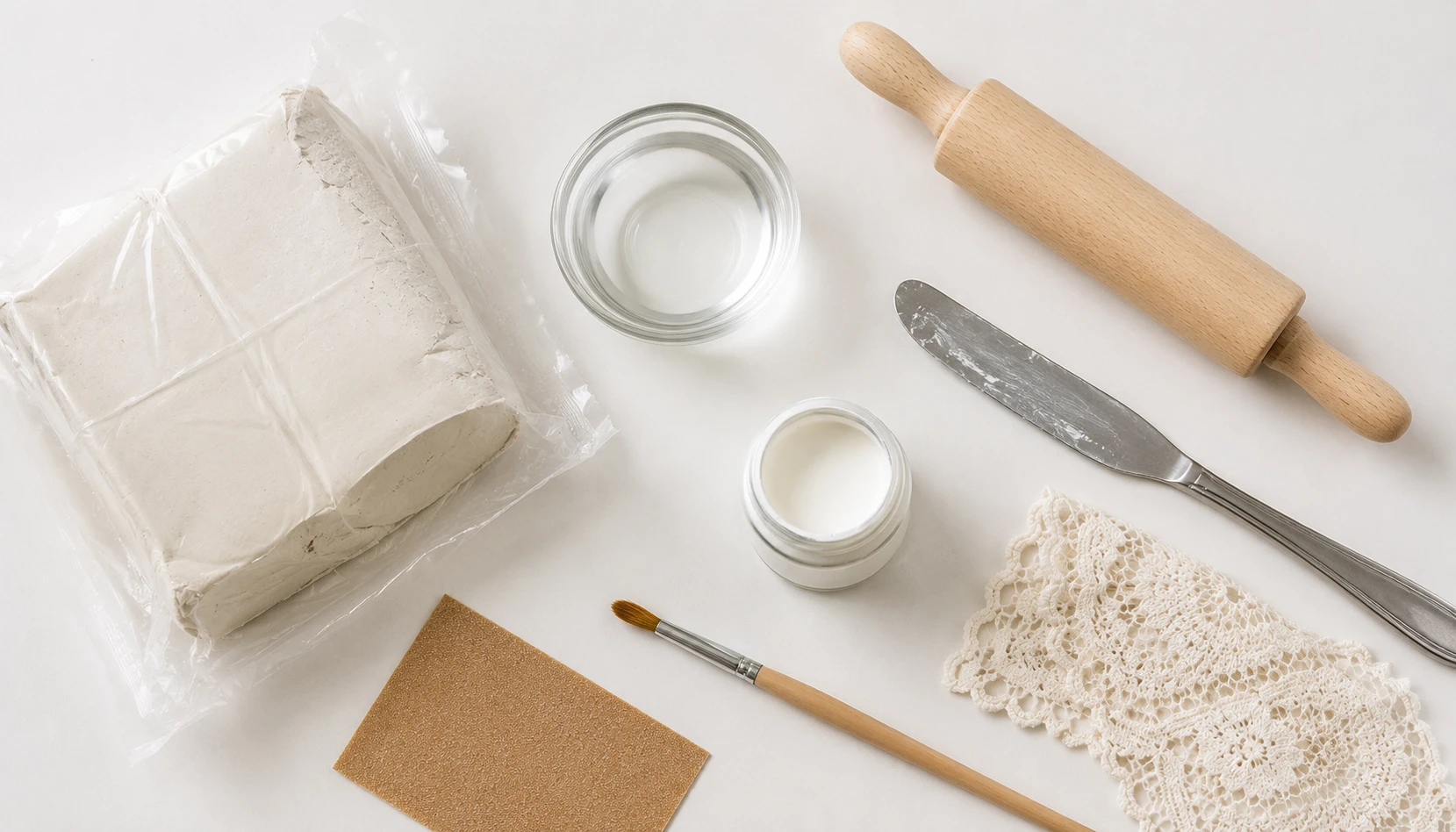 Flat lay of air dry clay supplies including a clay block, rolling pin, small bowl of water, and basic sculpting tools